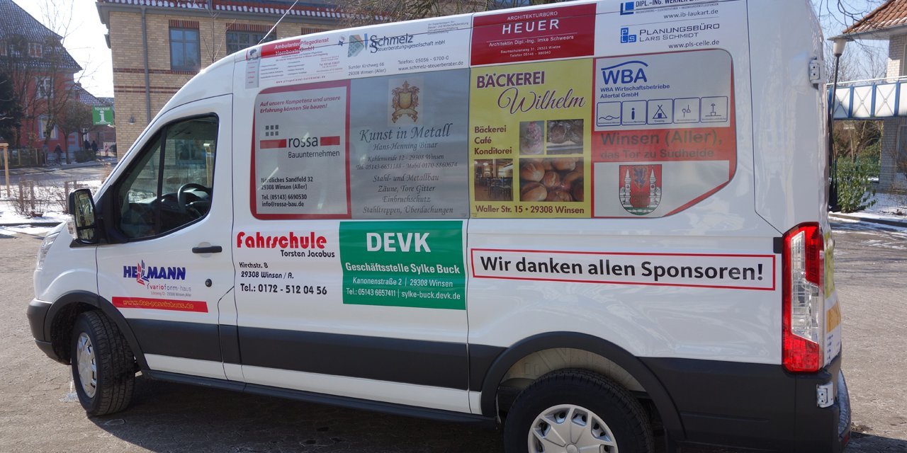 Weißer Kleinbus, Seitenansicht von rechts, auf einem Parkplatz, Bäume und Gebäude im Hintergrund.