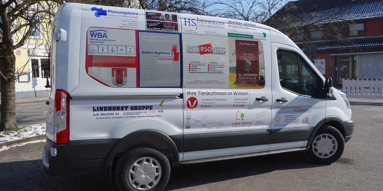 Weißer Kleinbus mit Werbeaufschriften, Seitenansicht von links, auf einem Parkplatz, Bäume und Gebäude im Hintergrund.
