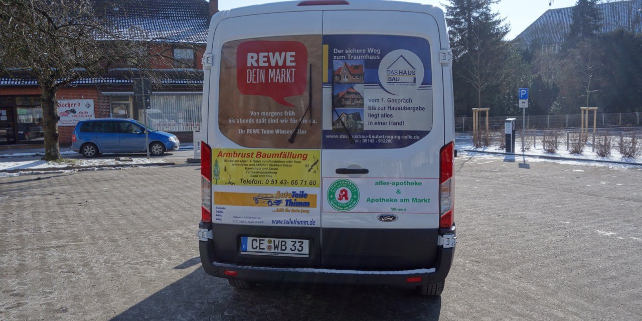 Weißer Kleinbus, Rückansicht, auf einem Parkplatz mit Bäumen und Gebäuden im Hintergrund.